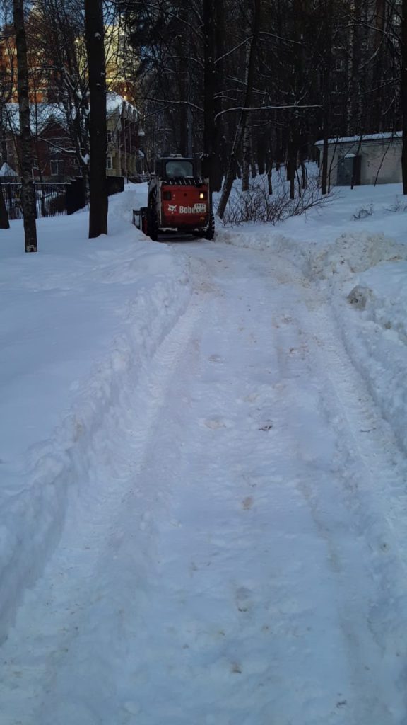 Пешеходная дорожка во время очистки снега в сквере. Снег чистит небольшой трактор оранжевого цвета. 