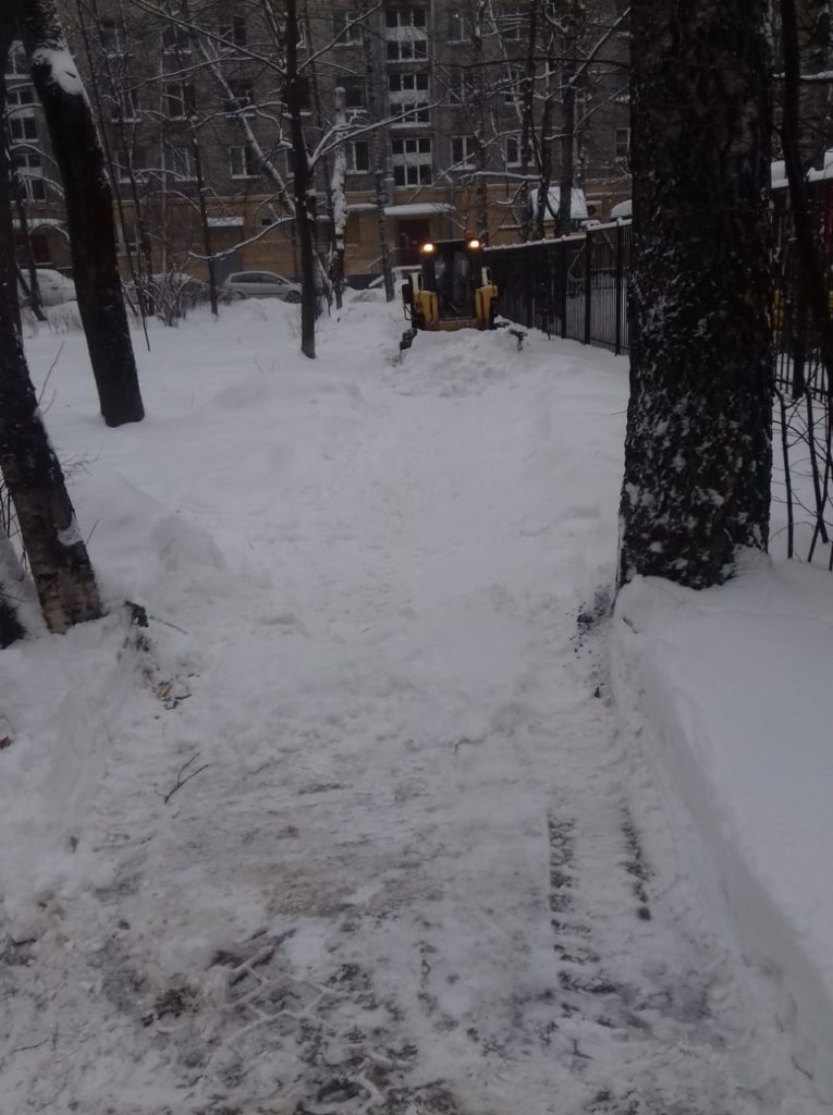 Пешеходная дорожка во время очистки снега в сквере. Снег чистит небольшой трактор оранжевого цвета. 