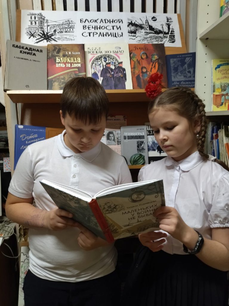 Дети рассматривают книгу "Маленьких у войны не бывает" на фоне книжных полок. 