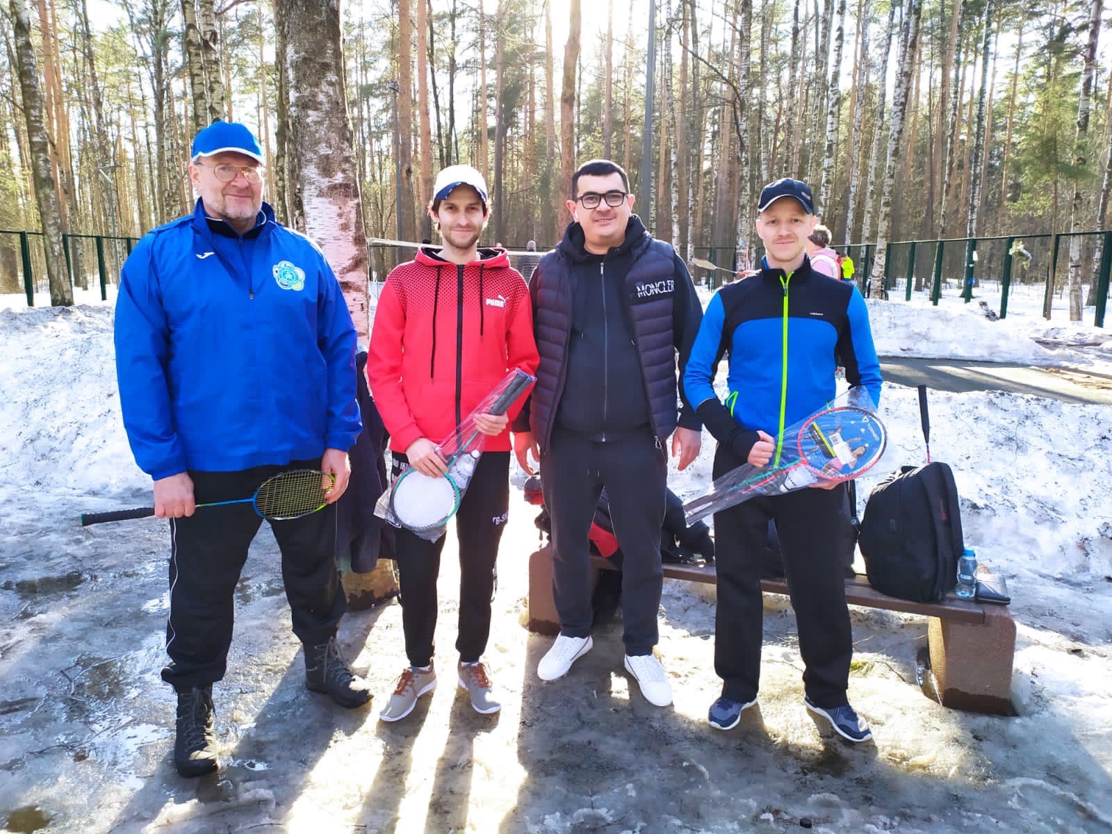 Глава Администрации МО Светлановское Сергей Кузьмин вручает участникам соревнований по бадминтону спортивный инвентарь. На фоне деревьев и снега стоят 4 человека в спортивных костюмах.