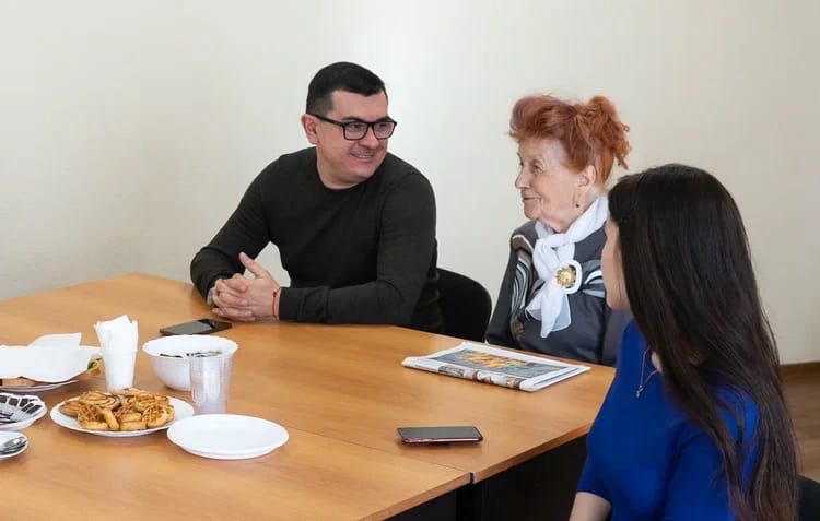 Глава МА МО Светлановское С.С. Кузьмин беседует с женщиной - узником фашистских концлагерей, сидя за столом.