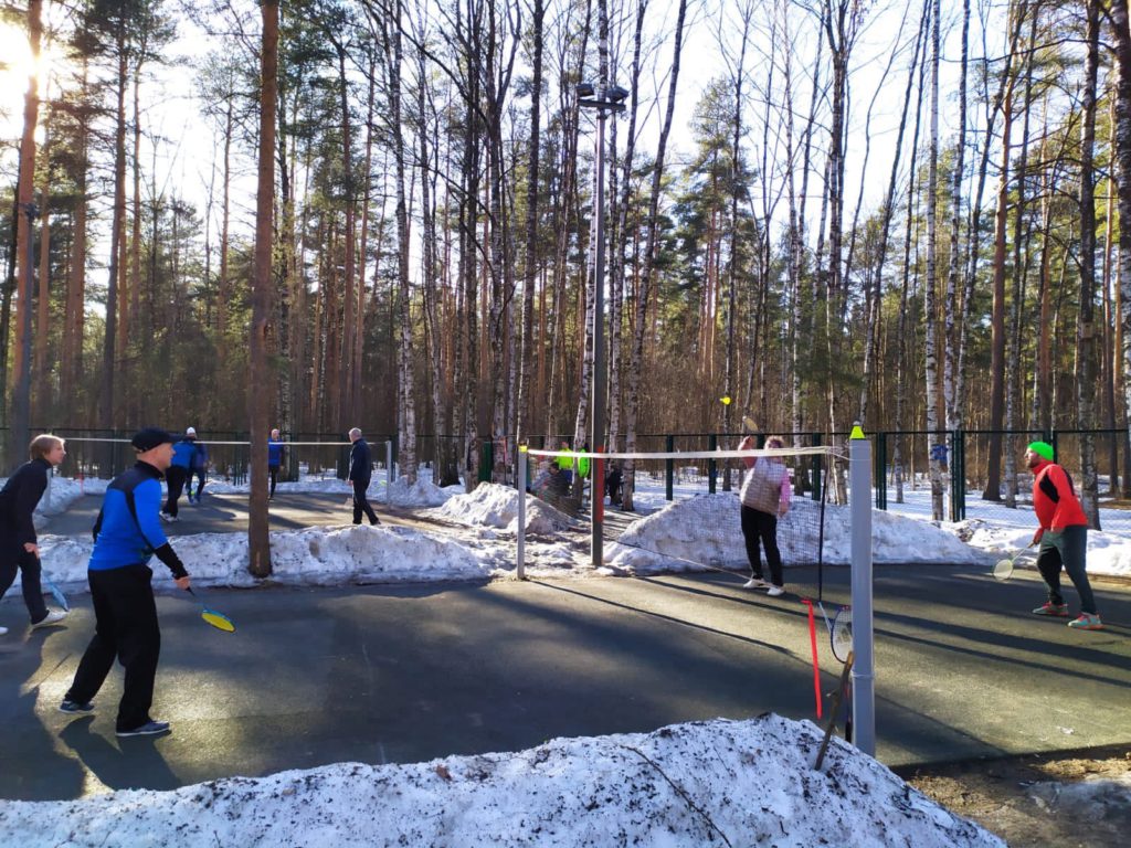 4 участника (4 человека в спортивных костюмах) соревнований играют в бадминтон. На фоне - деревья и снег. 