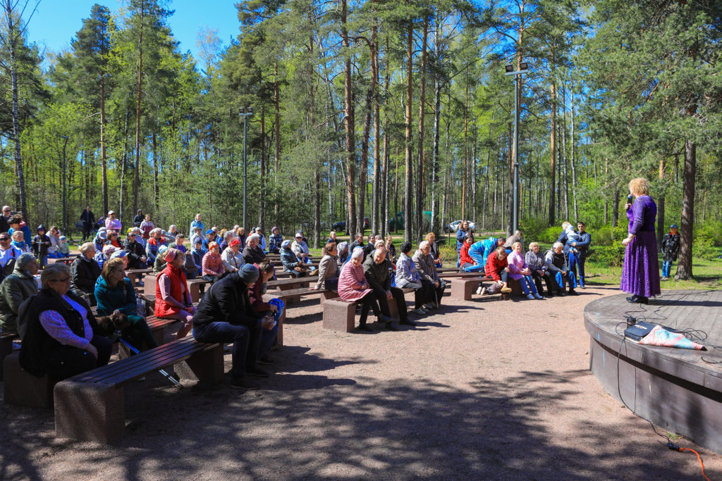 Жители МО Светлановское сидят на скамейках при прослушивании концерта духового оркестра в парке Сосновка.