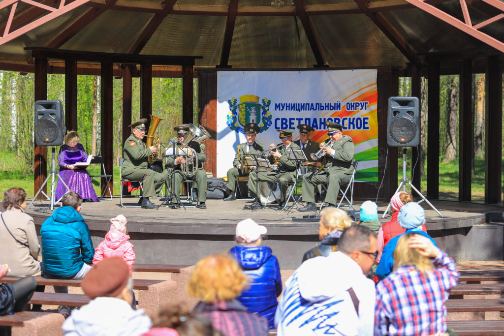 Музыканты духового оркестра (мужчины в военной форме) играют на духовых инструментах на сцене в парке Сосновка. На переднем плане - на скамейках сидят жители МО Светлановское и слушают выступление музыкантов. 