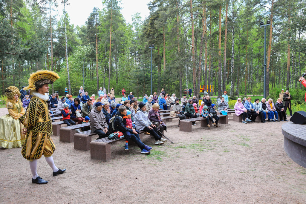 Концерт в парке Сосновка на летней эстраде на скамейках сидят жители и аплодируют. На фоне - зеленый лес. Сбоку от людей стоят аниматоры в пышных желтых костюмах под старину. 