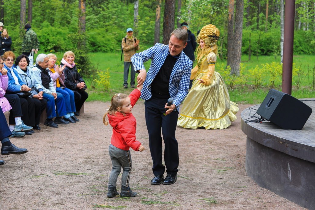 Концерт в парке Сосновка на летней эстраде - певец танцует с девочкой, на фоне - на скамейках сидят жители и аплодируют. На фоне - зеленый лес