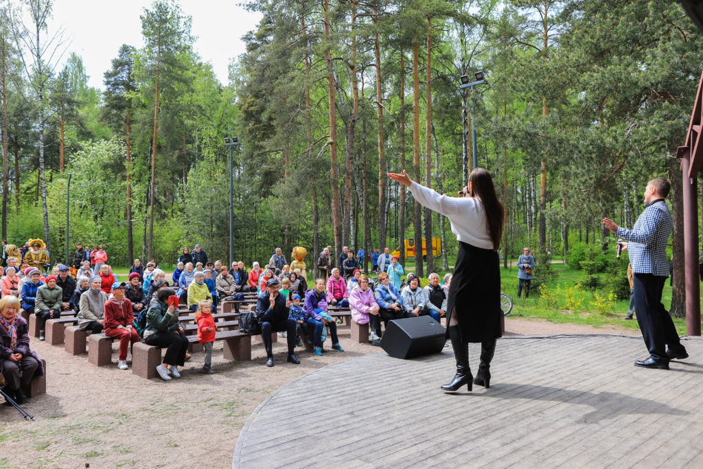 Концерт в парке Сосновка на летней эстраде - на сцене выступают певцы, на фоне - на скамейках сидят жители и аплодируют. На фоне - зеленый лес