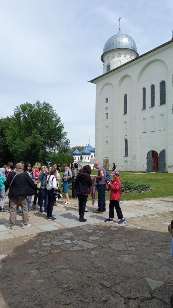 Люди на экскурсии в Великом Новгороде на фоне белокаменной церкви с серебристыми и голубыми куполами. 