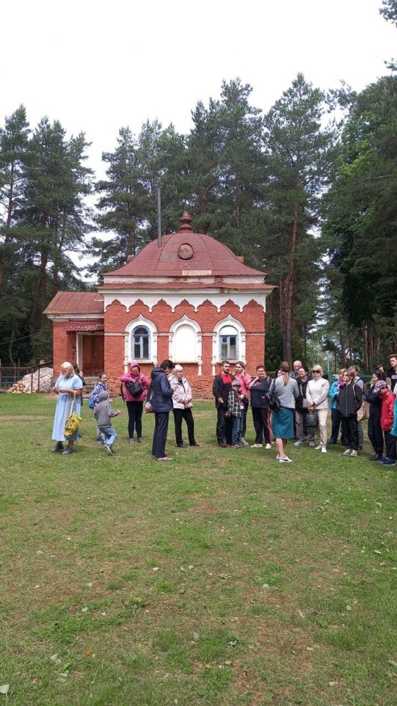 Люди на экскурсии в Великом Новгороде стоят на фоне здания из красного кирпича и зеленого хвойного леса. 
