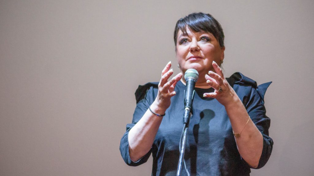 Женщина в темно-сером платье стоит перед микрофоном с поднятыми на уровне шеи руками. 