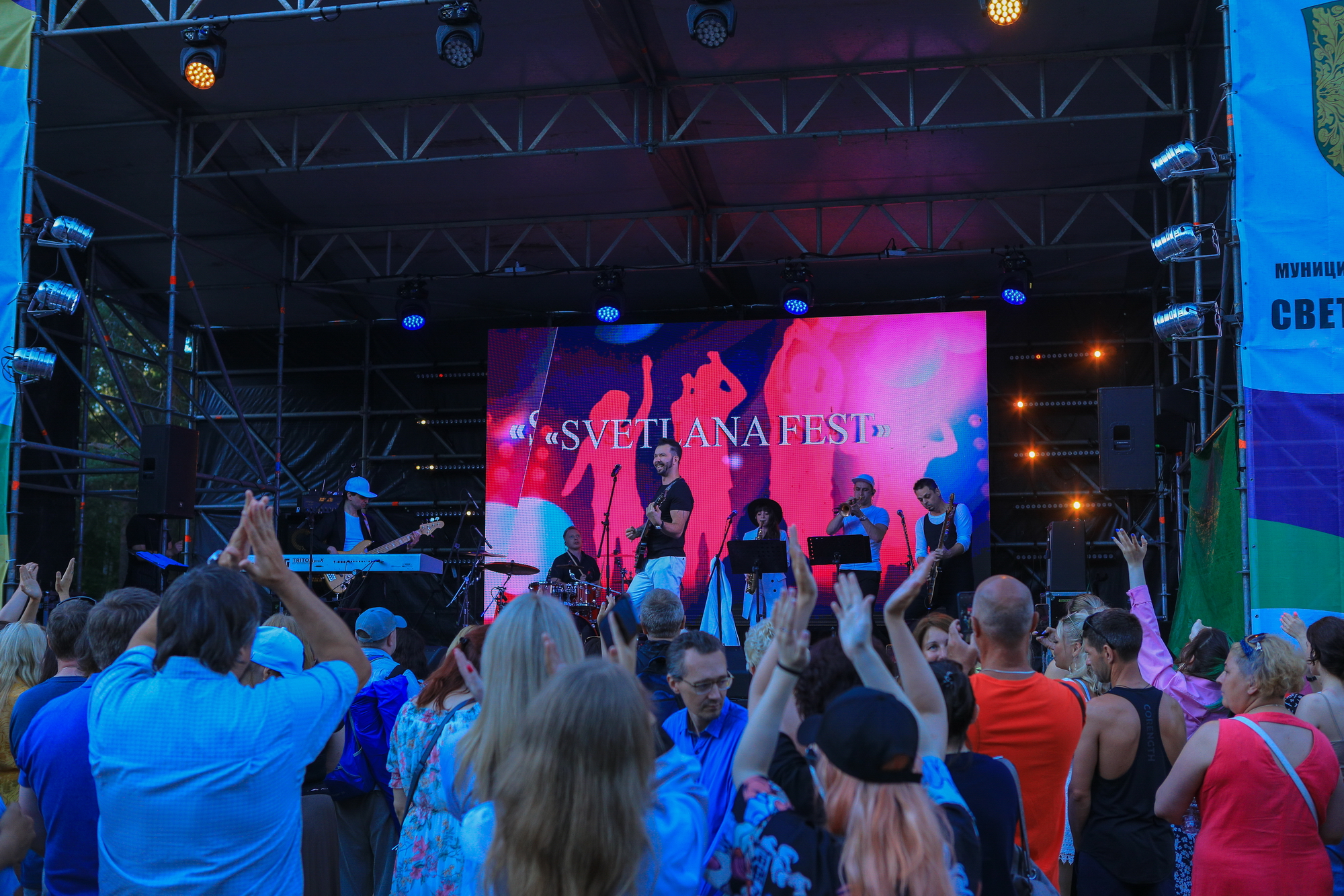 Артист Денис Клявер выступает на сцене в музыкантами. На фоне - люди подняли руки вверх и аплодируют.