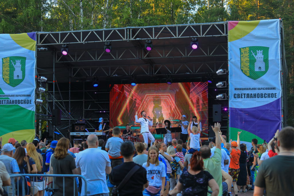 Артист Денис Клявер выступает на сцене с музыкантами. На фоне - люди стоят, танцуют и слушают выступление артиста. 
