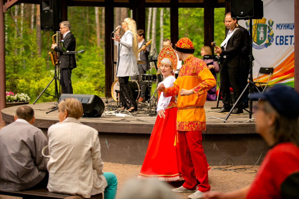 Аниматоры (парень и девушка) в русских народных костюмах танцуют на фоне играющих музыкантов. 