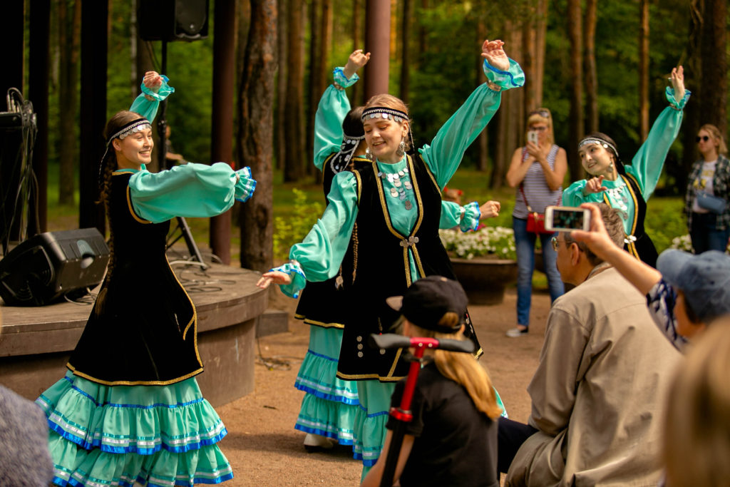 Девушки в башкирских народных костюмах танцуют национальный башкирский танец. На фоне - люди (зрители) и деревья.