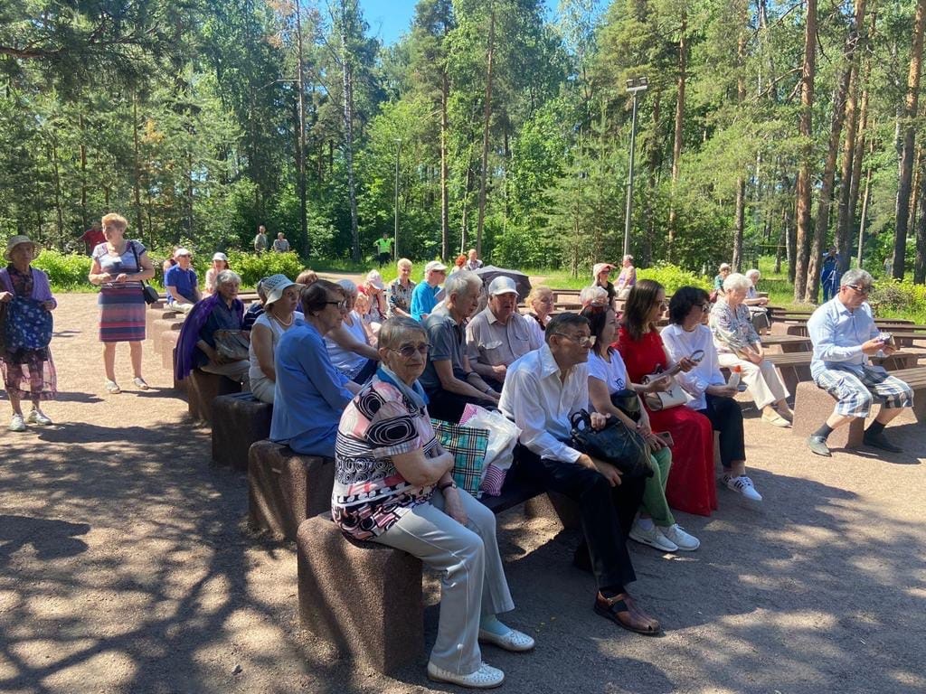Зрители наблюдают за выступлением духового оркестра в парке Сосновка на летней эстраде. На фоне - зеленые лес и трава. 