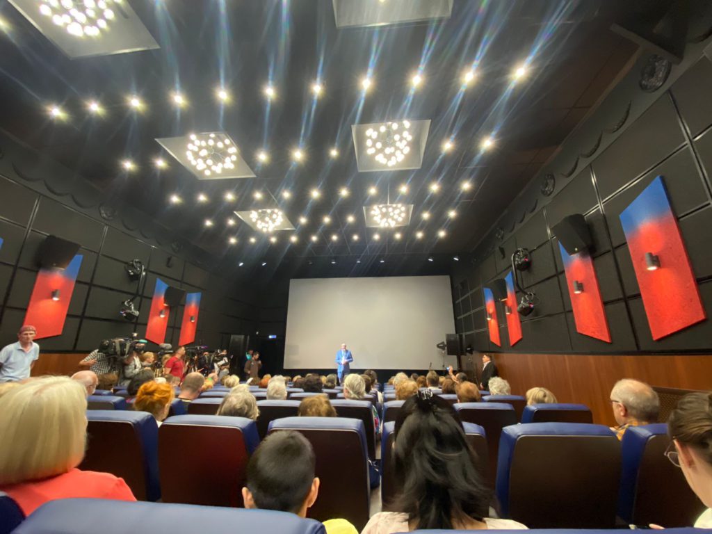Кинозал, люди сидят на креслах, на сцене выступает мужчина в синем костюме. 