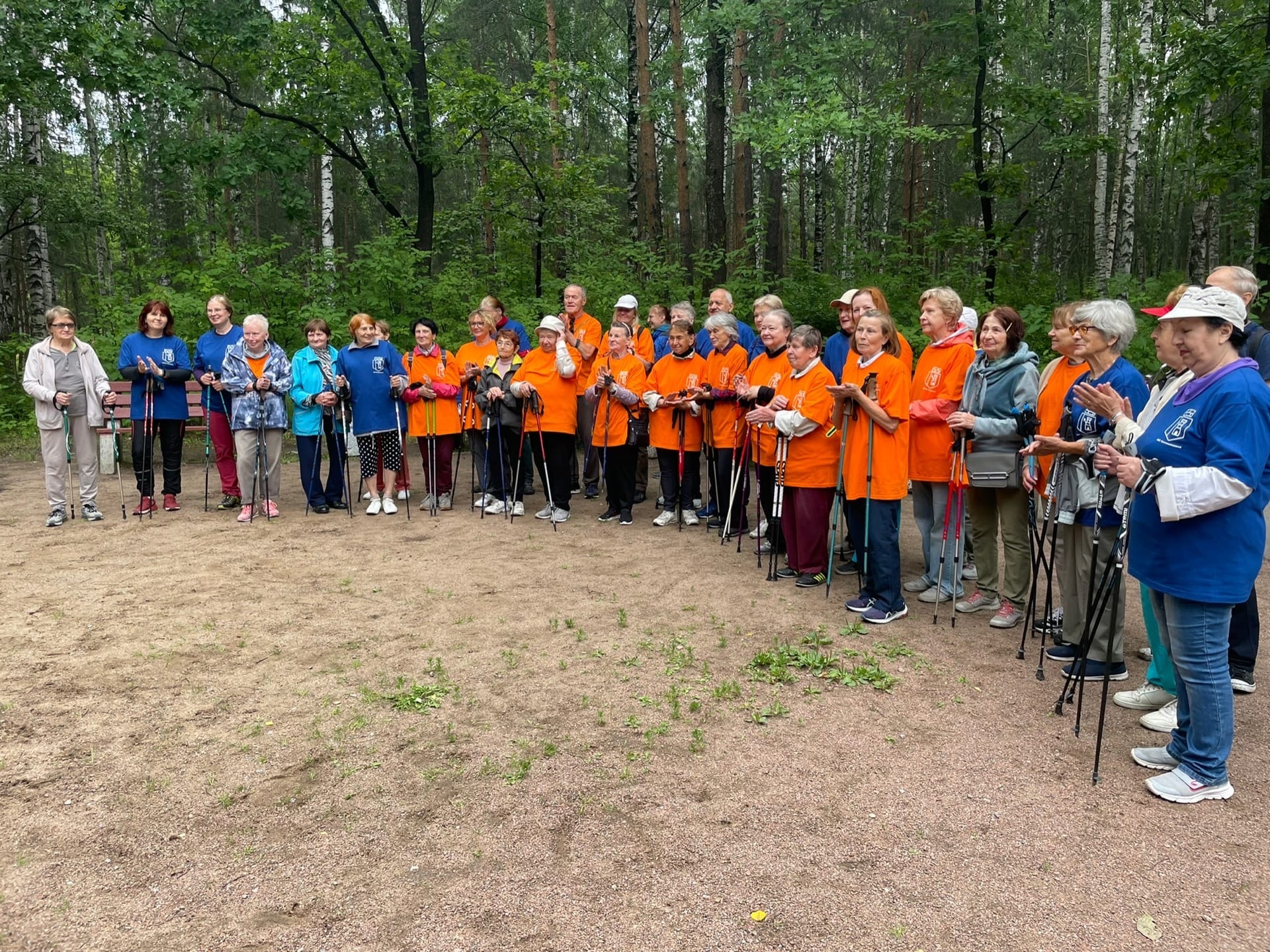 Группа жителей МО Светлановское в оранжевых и синих футболках с лыжными палками на занятии на скандинавской ходьбе.