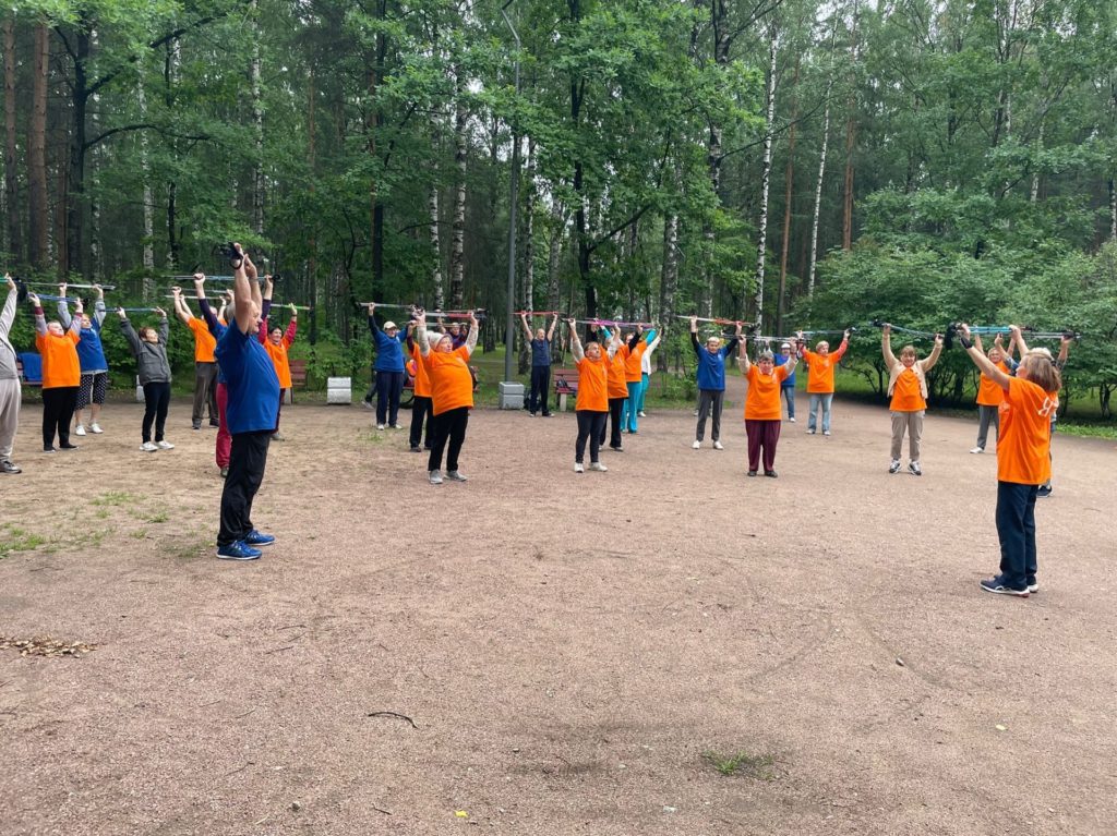 Группа жителей МО Светлановское в оранжевых и синих футболках делают разминку в рамках мастер-класса по скандинавской ходьбе. 