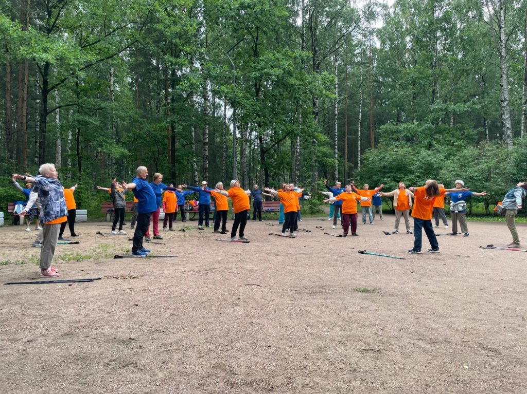 На фото: группа жителей МО Светлановское в оранжевых и синих футболках делает разминку в рамках мастер-класса по скандинавской ходьбе.